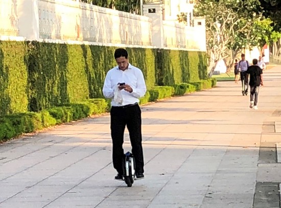 スマホ画面を見ながら川沿いの歩道を電動一輪車で走るビジネスマン、その後ろには電動キックスクーターが見える （筆者撮影）